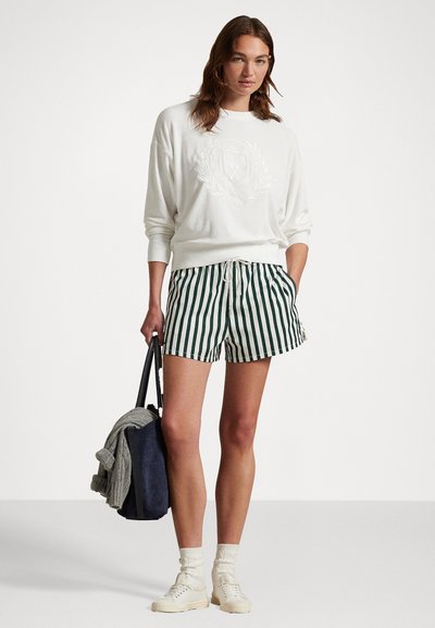 Hvit hettegenser med preget design, kombinert med grønne og hvite stripete shorts. Modellen holder en marineblå veske og har på seg hvite sneakers.
