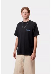 Camiseta de algodón negra con mangas cortas, que presenta un logo azul de "Carhartt WIP" en el área del pecho izquierdo. Combinada con pantalones de color claro.