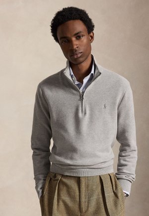 Jeune homme aux cheveux courts et bouclés portant un pull gris clair à fermeture éclair sur une chemise rayée et un pantalon marron à carreaux, les mains dans les poches.