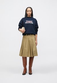 Φούτερ ναυτικού με λευκό λογότυπο «TOMMY HILFIGER», χακί πλισέ φούστα, καφέ μπότες μέχρι το γόνατο, κρατώντας μια μικρή μπεζ τσάντα.