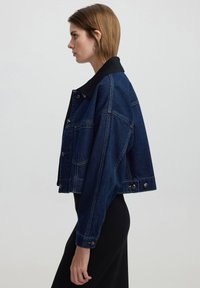 Chaqueta de denim en azul oscuro con un collar negro, que presenta un diseño corto, grandes bolsillos frontales y herrajes de botón plateados.