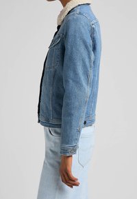 Džínová bunda ve světlemodré barvě s límcem z sherpy, obsahující boční kapsy, zapínání na knoflíky a viditelné švy.