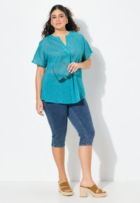 Turkos kortärmad tunika med V-ringning, matchad med mörka denimcapris och beige kilklacksandaler. Tyget har ett vågigt mönster.