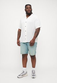 White short-sleeve button-up shirt with black buttons, paired with light blue denim shorts. Model wears gray sneakers and white socks.