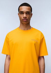 Žluté bavlněné tričko s krátkým rukávem, žebrovaným kulatým výstřihem a malým vyšitým logem na levé části hrudi. Hladká textura.