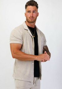 Kortärmad, ljusbeige knapp-up skjorta i ett strukturerat tyg, kombinerad med en svart t-shirt. Modellen har ett neutralt uttryck och en muskulös kropp.