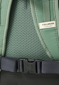 Retro in verde della rete di uno zaino con cinture e fibbia blu scuro. Presenta l'etichetta del marchio "Fjällräven" in testo bianco. Superficie texturizzata.