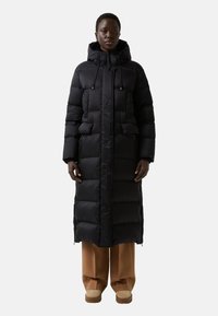 Long manteau en duvet noir avec capuche, comportant des sections matelassées, une fermeture éclair à l'avant et des poches latérales. Porté par-dessus un pantalon large marron et des chaussures.