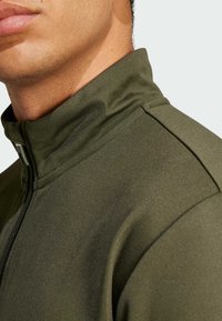 Olive green zip-up jacket with a high collar, textured fabric, and minimalistic design. Features a sleek zipper along the front.