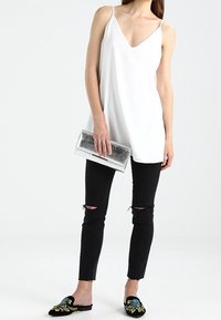 Kvinna i ärmlös vit topp, svarta ripped skinny jeans, svarta blommiga slip-on skor, håller i en silverfärgad metallisk clutch.