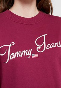 Burgundy collegepaita, jossa on ribbireunus ja valkoisella kirjailtu teksti "Tommy Jeans" sekä pieni amerikkalaisen lipun koriste.
