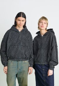 Black hooded jackets with textured patterns. Features zip closures, elastic cuffs, and hem. Paired with light and dark denim trousers.