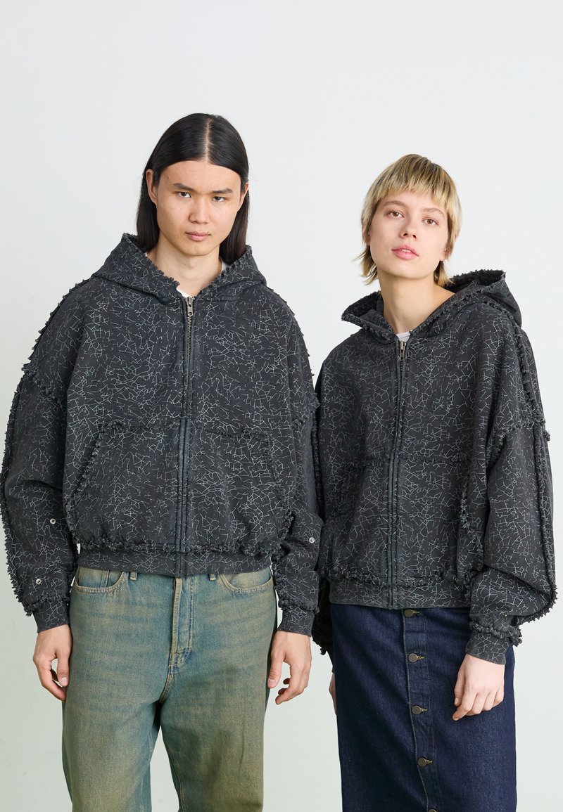 Black hooded jackets with textured patterns. Features zip closures, elastic cuffs, and hem. Paired with light and dark denim trousers.