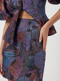 Σύνολο δύο κομματιών με μαύρη βάση και φωτεινά πολύχρωμα μεταλλικά μοτίβα. Το τοπ έχει φουσκωτά μανίκια και η φούστα έχει διακοσμητικά κουμπιά.