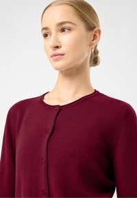Burgundský cardigan z měkkého pleteného materiálu, který má kulatý výstřih, zapínání na knoflíky a dlouhé rukávy. Texturovaná látka s jemným elastickým efektem.