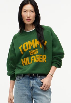Femme aux longs cheveux foncés portant un sweat-shirt vert Tommy Hilfiger et un jean bleu, debout avec une main dans la poche.