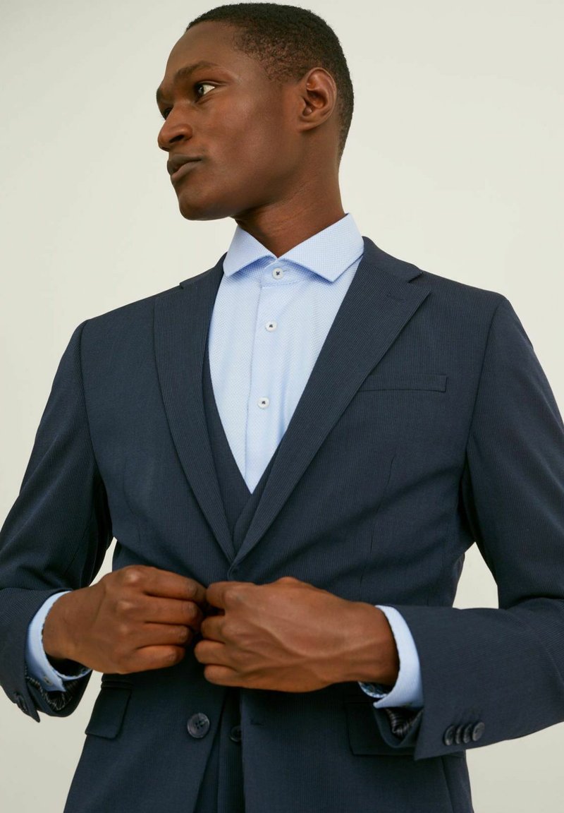 Man fäster knappen på en mörkblå kostymjacka över en ljusblå skjorta mot en neutral bakgrund.