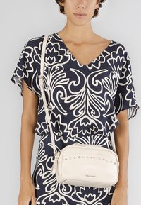 Borsa a spalla crema con borchie decorative sul davanti, realizzata in pelle con una texture liscia, abbinata a una blusa con stampa floreale blu navy.