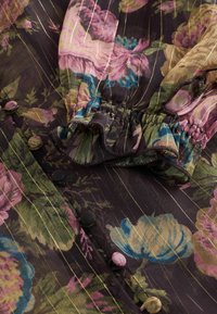 Permatomas gėlių bluzonas su tamsiu fonu, sudėtingomis aukso juostelėmis, susiraukšliu kaklu ir daugiaspalviais sagomis. Pasižymi sluoksniuota tekstūra.