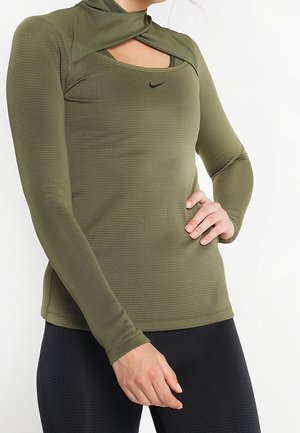 Vrouw draagt een olijfgroen sporttopje met lange mouwen en een uitgesneden halslijn en zwarte leggings, hand op heup tegen een witte achtergrond.
