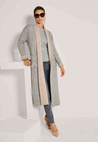 Frau in einem langen grauen Cardigan mit beigem Saum, mit verzierten Taschen, grauem Oberteil, dunklen Jeans, beigen Absätzen, großen Sonnenbrillen und langen Ohrringen.