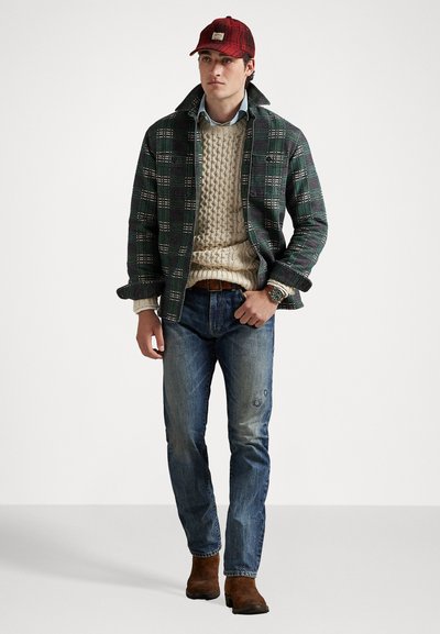 Muž v červenej kockovanej čiapke, zelenej kockovanej bunde, béžovom pletenom svetri, modrých džínsoch, hnedých čižmách a s hodinkami, stojí na pozadí bez vzoru.