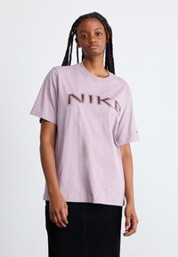Camiseta de algodón de color púrpura claro con mangas cortas, que presenta un logo "NIKE" en negrita y color rojo oscuro en la parte frontal. Diseño de cuello redondo simple.