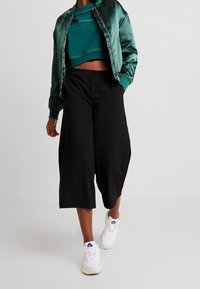 Grön satinbombarjacka över en kort teal sweatshirt och svarta culottes, tillsammans med vita sneakers. Släta texturer och avslappnad passform.