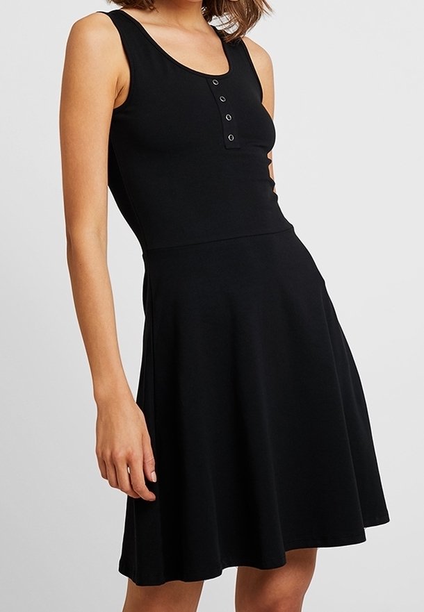 Femme portant une robe noire sans manches avec un col rond boutonné et une jupe évasée, debout devant un fond clair uni.