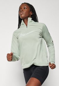 Sudadera atlética verde claro con cremallera, presenta un cuello alto y mangas largas, confeccionada con un material suave y flexible que incluye un logo reflectante.