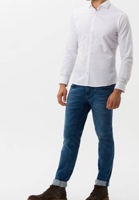 Bílá košile s dlouhým rukávem na knoflíky, modré džínové džíny s ohrnutými lemy a hnědé kotníkové boty s měkkou texturou a přiléhavým střihem.