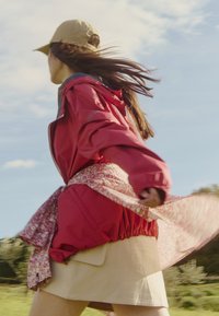 Rode waterdichte jas met elastische boorden, bloemenpatroon stof omslag en beige rok. In de wind waaien haar tegen een blauwe lucht achtergrond.