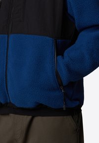 Polarowa kurtka z niebieską dolną częścią i czarną górną częścią. Posiada kieszenie na zamek oraz regulowany sznurek w dolnej części. Miękka faktura.