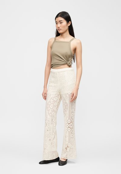 Γυναίκα με μακριά σκούρα μαλλιά, φορώντας ένα ελιάς τοπ δεμένο κόμπο, παντελόνι εκρού με φαρδιές δαντέλες και μαύρα επίπεδα παπούτσια, στέκεται μπροστά σε απλό φόντο.