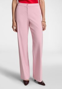 Pantaloni modello tailored rosa chiaro con gamba dritta, frontale piatto e chiusura a bottone discreta, abbinati a scarpe nere con punta affusolata.