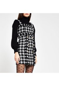 Schwarz-weißer Hahnentrittkleid mit strukturiertem Design, ausgestattet mit goldenen Knöpfen und einem strukturierten Stoff, kombiniert mit einem schwarzen Rollkragenpullover.