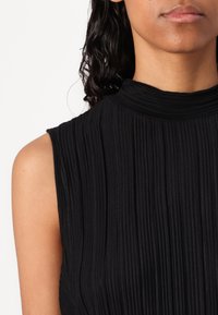 Kobieta o średnim odcieniu skóry, ubrana w czarny, bezrękawnikowy top z prążkowanego materiału o wysokim dekolcie, odsłaniająca ramię i dolną część twarzy z zamkniętymi ustami.