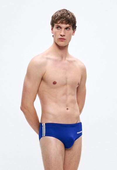 Blaue Badehose mit weißen Seitenstreifen, elastischem Material, figurbetontem Design und einem Logo auf der Vorderseite. Modell steht in neutraler Haltung.