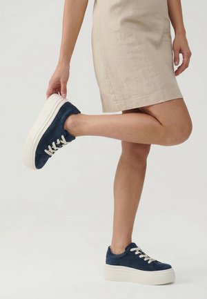 Persoon draagt een beige rok en houdt één voet omhoog, waarbij donkerblauwe platformsneakers met witte veters en dikke witte zolen worden getoond.