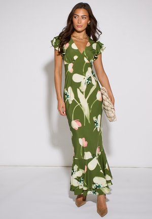 Woman wearing a green floral maxi dress with ruffled sleeves and hem, holding a woven clutch, standing against a plain background.