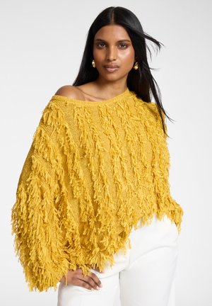 Vrouw draagt een gele, gestructureerde gebreide trui met franjes, waarbij één schouder bloot is, en witte broek, staand tegen een effen achtergrond.