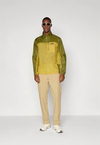 Muška pulover jakna u maslinastoj zelenoj i žutoj boji, izrađena od laganog materijala. Ima četvrtasti patent zatvarač, duge rukave i bočne džepove.