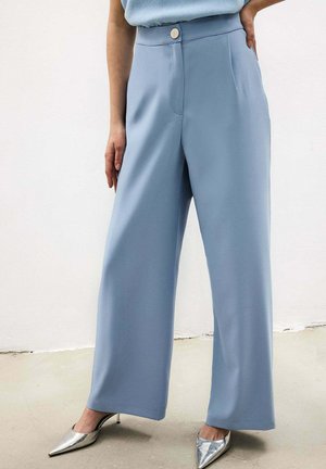 Osoba v svetlomodrých širokých nohaviciach a strieborných vysokých podpätkoch so zašpicatenou špičkou, stojaca s jednou rukou v vrecku.