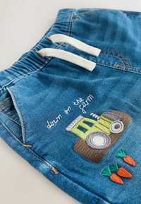 Sinivalkoiset farkkushortsit, joissa valkoiset kiristysnauhat, koristeltu vihreällä traktorilla ja porkkanalla kirjailtuna sekä tekstillä "down on the farm".