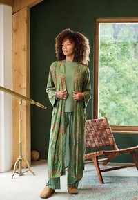 Lange groene kimono met een patroon en brede mouwen over een groene top en gestreepte broek. Zachte slippers; natuurlijk hout en groen in de achtergrond.