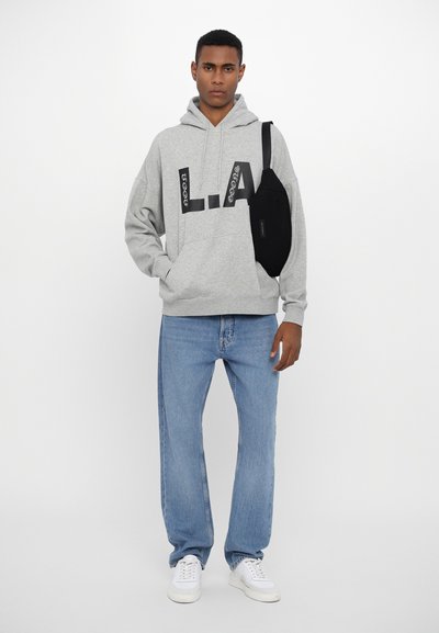 Haut à capuche gris avec impression "L.A." en noir, associé à un jean bleu clair et des baskets blanches, avec un sac banane noir porté en travers de l'épaule.