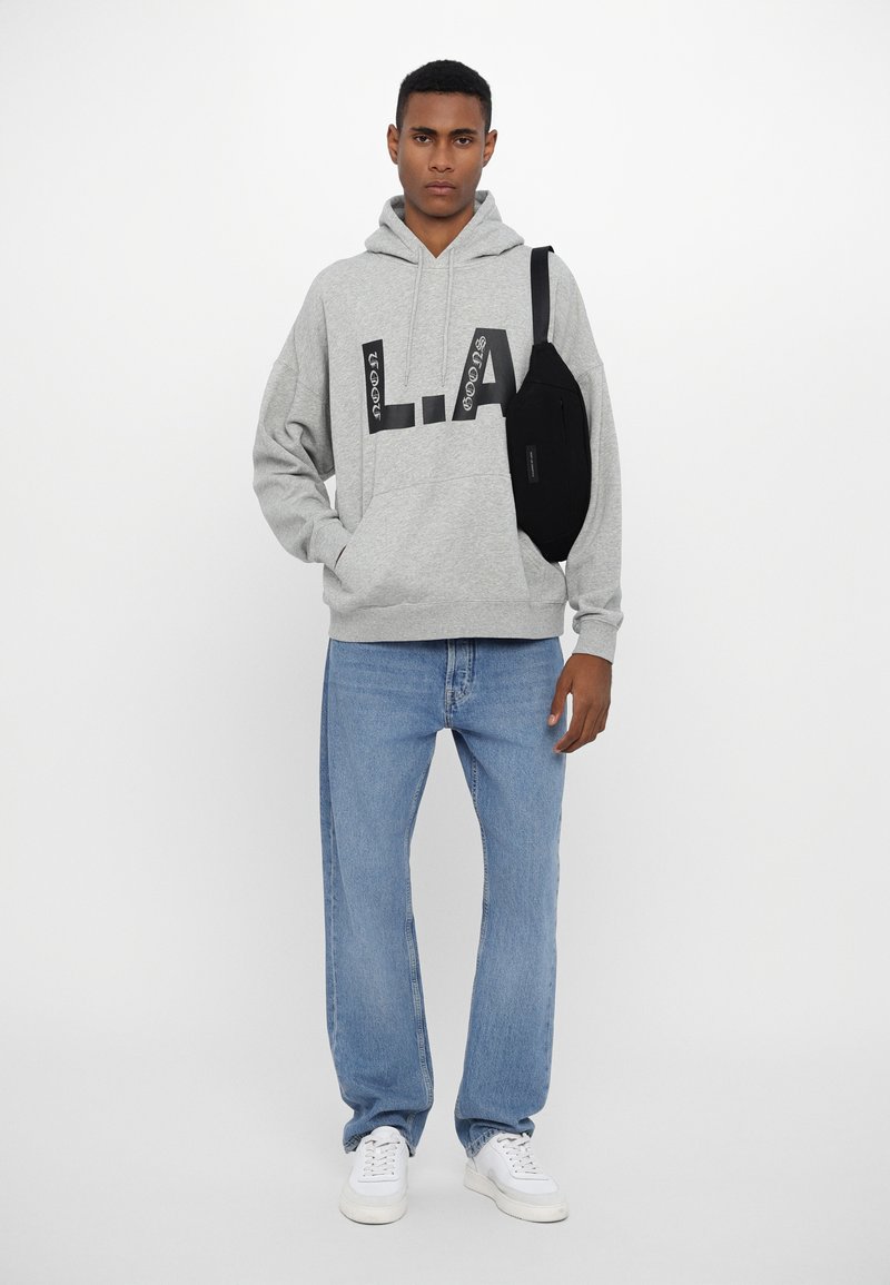 Grau Hoodie mit schwarzem "L.A."-Druck, kombiniert mit hellblauen Jeans und weißen Sneakers, dazu eine schwarze Umhängetasche, die über die Schulter getragen wird.