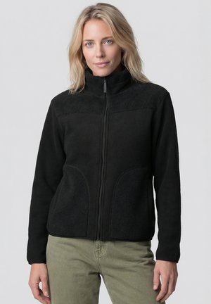 Frau mit blonden Haaren, die eine schwarze Fleecejacke mit Reißverschluss und olivgrüne Hose trägt, vor einem schlichten hellen Hintergrund stehend.