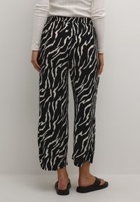Personne portant un pantalon large à motif abstrait noir et blanc, un haut à manches longues blanc et des sandales noires, debout devant un mur uni.