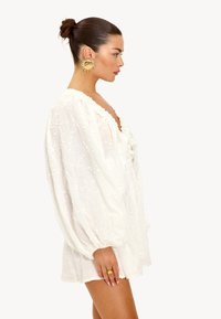 Witte geborduurde jurk met lange pofmouwen, diepe halslijn en gerimpelde afwerking. Geaccentueerd met grote ronde gouden oorbellen en een ring.